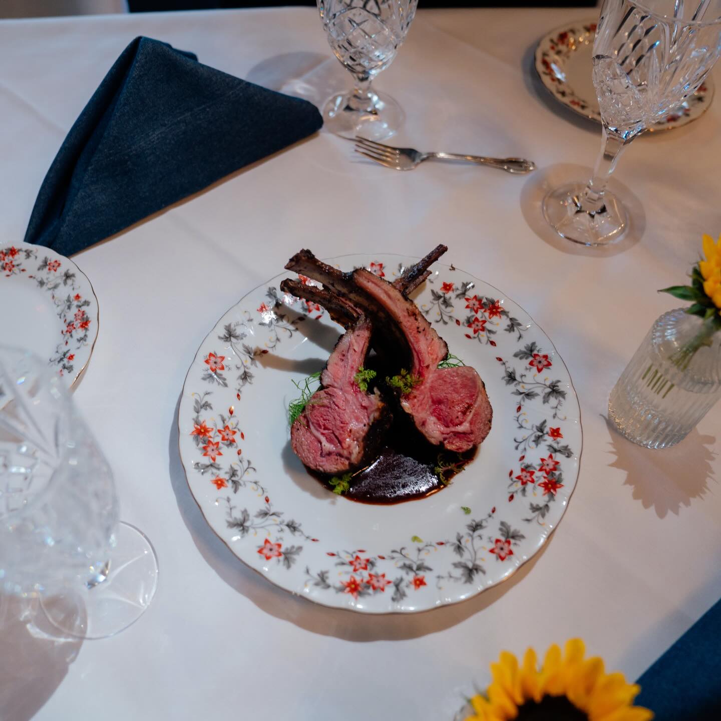 Two pink lamb chops on floral china beside a navy napkin and a sunflower